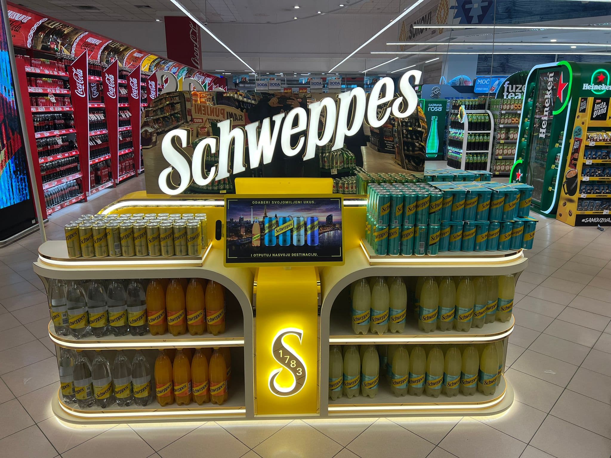 Illuminated Schweppes branded POS island in a retail store, with product shelving and an interactive display screen